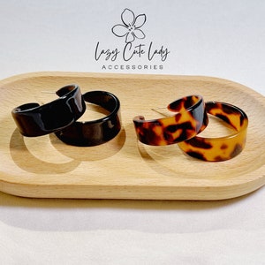 May include: Two pairs of hoop earrings displayed on a wooden tray. One pair is black and the other is brown with a tortoiseshell pattern. The earrings are made of plastic and have a shiny finish. The tray is oval shaped and is light brown in colour. The text "Lazy Cute Lady Accessories" is printed on the tray.