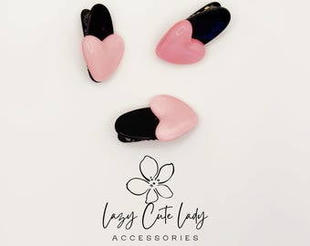 Puede incluir: Tres pinzas para el pelo en forma de corazón negro y rosa. Las pinzas tienen un acabado brillante y un diseño de corazón rosa claro. La marca "Lazy Cute Lady Accessories" está escrita en cursiva debajo.