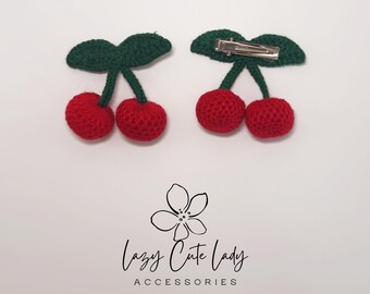 Puede incluir: Dos pinzas para el pelo de cerezas de ganchillo. Cada pinza presenta dos cerezas rojas con tallos y hojas verdes. Una pinza tiene una horquilla plateada en la parte posterior. El texto "Lazy Cute Lady ACCESSORIES" está debajo.