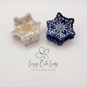 May include: Two snowflake-shaped hair clips. One is white with gold accents and clear rhinestones, the other is navy blue with white accents and clear rhinestones. The text "Lazy Cute Lady Accessories" is below the clips.