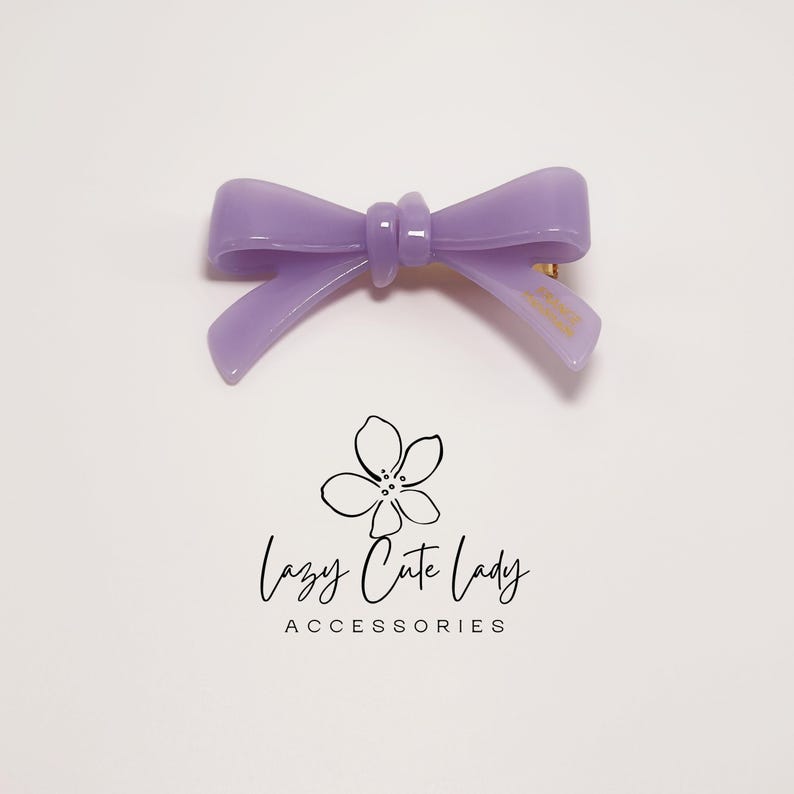 Pu&ograve; includere: Un fermaglio per capelli a forma di fiocco color lavanda. Il fiocco &egrave; realizzato in materiale lucido e ha una forma classica. Il fermaglio &egrave; di colore viola chiaro e ha una chiusura dorata. Il testo "Lazy Cute Lady Accessories" &egrave; sotto.