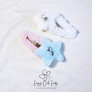 May include: Two fuzzy star-shaped hair clips with rhinestones. The clips are pink and blue with white fur.