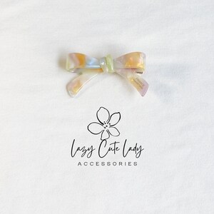 Pu&ograve; includere: Un fermaglio per capelli a forma di fiocco perlato con un centro verde chiaro e un gradiente oro e viola. La clip &egrave; etichettata "FRANCE handmade". Il logo "Lazy Cute Lady Accessories" &egrave; sotto.