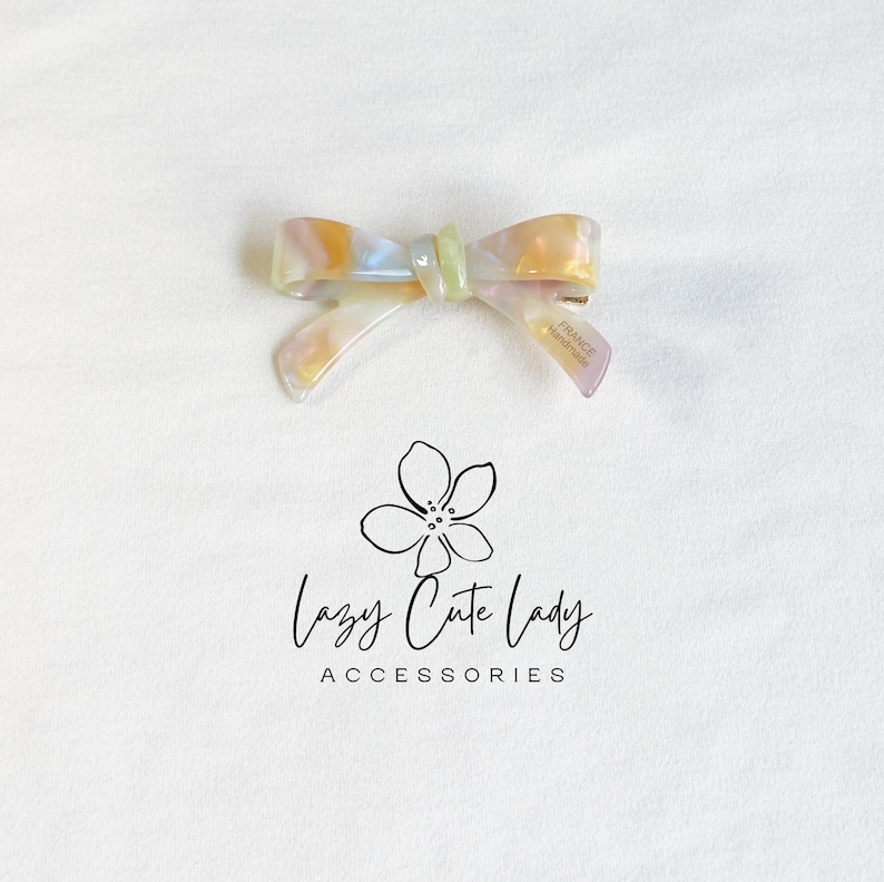 Pu&ograve; includere: Un fermaglio per capelli a forma di fiocco perlato con un centro verde chiaro e un gradiente oro e viola. La clip &egrave; etichettata "FRANCE handmade". Il logo "Lazy Cute Lady Accessories" &egrave; sotto.