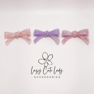 Pu&ograve; includere: Tre fermagli per capelli a forma di fiocco, nei toni del rosa e del viola. I fermagli hanno una finitura lucida e sono disposti su una superficie bianca. Il testo "Lazy Cute Lady Accessories" &egrave; sotto.