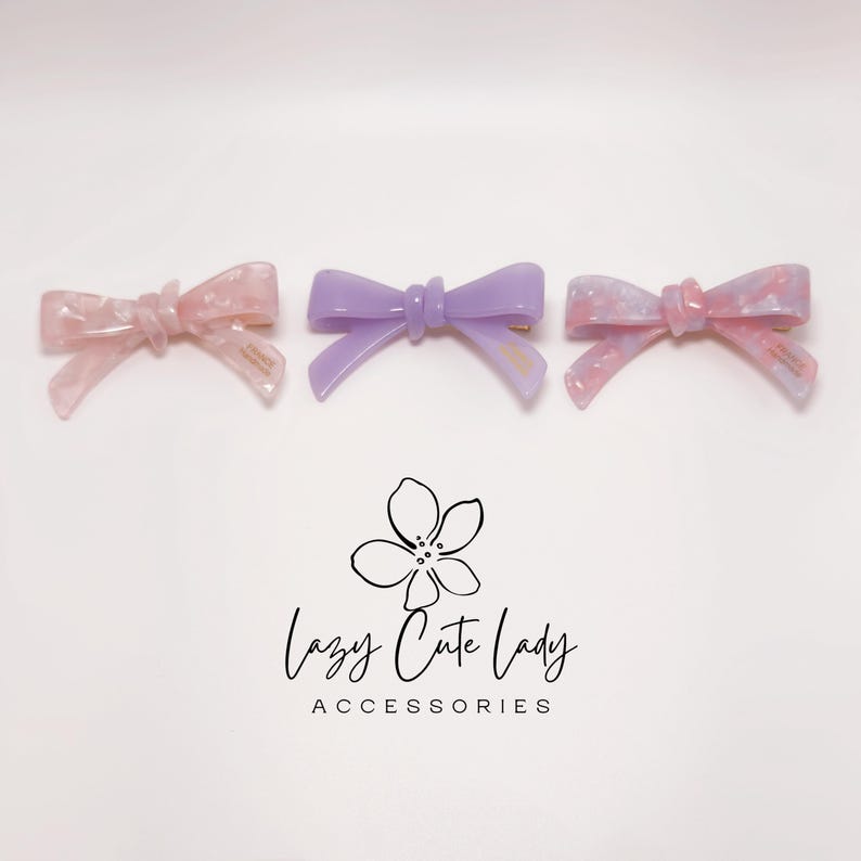 Pu&ograve; includere: Tre fermagli per capelli a forma di fiocco, nei toni del rosa e del viola. I fermagli hanno una finitura lucida e sono disposti su una superficie bianca. Il testo "Lazy Cute Lady Accessories" &egrave; sotto.