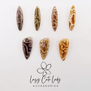 May include: Seven polished, oval-shaped nail art accessories in shades of brown, tan, and pink, with unique swirling patterns. The accessories are arranged on a white surface. The image includes the text "Lazy Cute Lady Accessories".