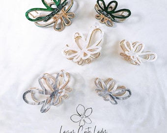May include: Assortment of six butterfly-shaped hair clips in green, white, and gray. The clips are made of plastic and have a gold metal clasp. The clips are arranged on a white surface. Lazy Cute Lady Accessories is printed below the clips.