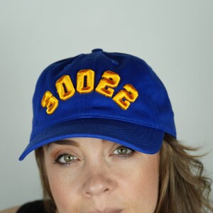 May include: A woman wearing a blue baseball cap with the text "2023" in yellow embroidered on the front. She has long brown hair and is wearing a black tank top.