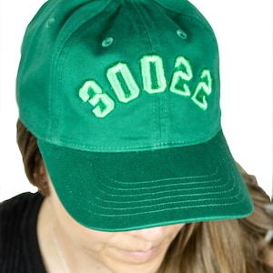 Op de afbeelding: Een groene baseballpet met het nummer "3002" in wit geborduurd op de voorkant.