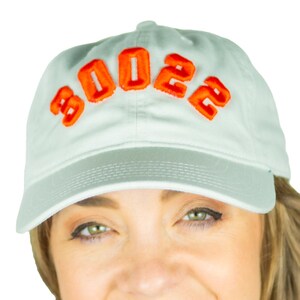 Può includere: Un berretto da baseball blu chiaro con la parola "2022" ricamata in arancione sul davanti.