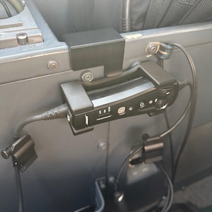 May include: A black Bose audio device is mounted on a grey surface with several black clips and wires. The device has buttons and switches, and the Bose logo is visible. The image is taken in a vehicle.
