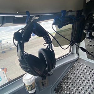 May include: A pair of black Bose aviation headsets hanging from a blue 3D-printed hook attached to the cockpit window frame. A white coffee cup with a green logo sits in a circular holder below the headset, near the aircraft control panel.