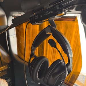 May include: A black aviation headset with cushioned ear cups and an adjustable boom microphone, suspended from a grey plastic mount attached to an overhead console. A black control module with buttons and a dial is secured to the mount above.