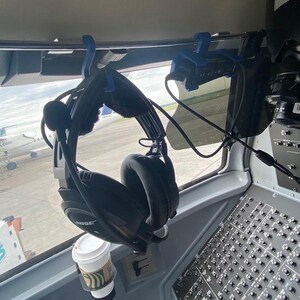May include: Black aviation headset with a microphone boom arm hanging from a blue plastic hook mounted on a metal bar inside an aircraft cockpit. A white paper coffee cup sits in a circular holder below the headset near a control panel.
