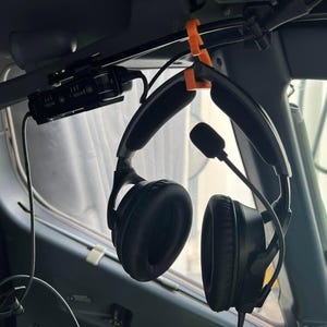 May include: Black aviation headset with padded ear cups and an adjustable microphone boom, suspended from a bright orange plastic hook attached to the overhead panel of an aircraft cockpit, featuring a black cable and a control module.
