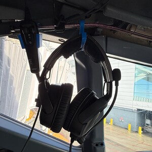May include: A black aviation headset with a microphone boom and a black handheld control unit, both suspended from blue plastic hooks mounted to the interior frame of an aircraft cockpit, with a view of an airport tarmac visible through the window.