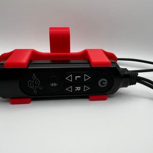 May include: A black electronic device with a red bracket. The device has several buttons with directional arrows and a power symbol. Cables extend from both ends. The device is on a white surface.