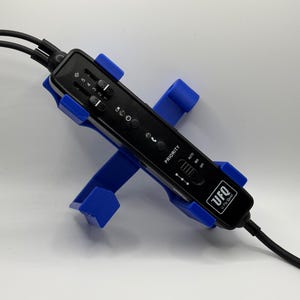 May include: A black electronic device with the text "UFO" and "U FQ" is held by a blue plastic stand. The device has multiple buttons and sliders, and is connected to black cables. The stand is in the shape of an X.