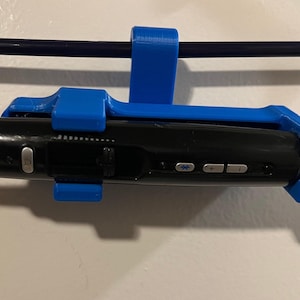 May include: A bright blue 3D-printed plastic holder securely gripping a black electronic device with a cord. The holder features a hook design for hanging on a rod, while the black device displays silver buttons, a dial, and a small blue icon.