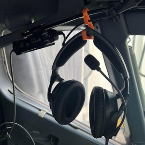 May include: A black aviation headset with padded ear cups and an adjustable microphone boom, suspended from a bright orange 3D-printed plastic hook attached to the overhead panel of an aircraft cockpit, featuring a coiled black connection cable.