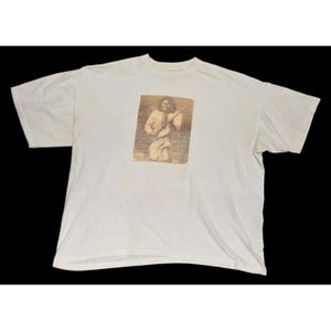 May include: Off-white t-shirt featuring a sepia-toned image of a Native American man kneeling and holding a weapon. The image is rectangular and centered on the front of the shirt. The shirt has short sleeves and a crew neck.