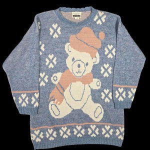 May include: A light blue knit sweater featuring a large teddy bear design with a pink hat and scarf. The sweater also has white snowflake patterns and a pink border at the hem and cuffs. The label reads "ADELEY KNITWEAR".