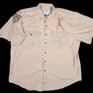 May include: A tan short-sleeved button-down shirt featuring two chest pockets, a collar, and dark buttons. The front displays the text "FIRST AIR COMMANDS GROUP" and a graphic of a winged figure. A shield-shaped patch is on the right sleeve.