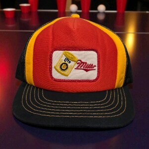 May include: A red, yellow, and black trucker hat with a black brim. The hat features a rectangular patch with a beer can graphic and the word "Miller" in red script. The hat has a mesh back.