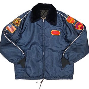 May include: Navy blue jacket with a black faux fur collar, white piping, and multiple patches. The jacket features a zipper closure, black cuffs, and a red and yellow name patch. The patches include an American flag and other emblems.