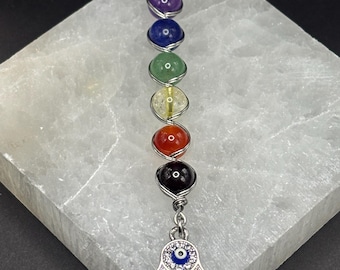 May include: A chakra-themed keychain featuring seven colorful gemstone beads, each wrapped in silver wire. A silver Hamsa hand charm with a blue eye and clear crystals dangles from the bottom. The keychain has a silver clasp.