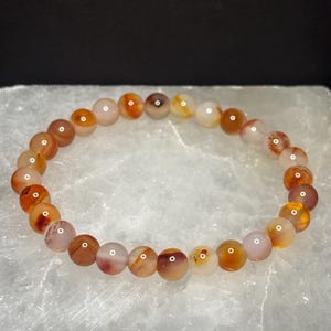 Puede incluir: Un brazalete de cuentas redondas y pulidas en tonos naranja, marrón y crema. Las cuentas están ensartadas para formar un brazalete circular. El brazalete se exhibe sobre una superficie blanca.