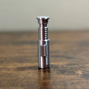 Puede incluir: Peque&ntilde;o objeto cil&iacute;ndrico de metal plateado con superficie texturizada. El objeto tiene una serie de crestas horizontales y una secci&oacute;n rectangular con l&iacute;neas verticales. La parte superior tiene un dise&ntilde;o circular.
