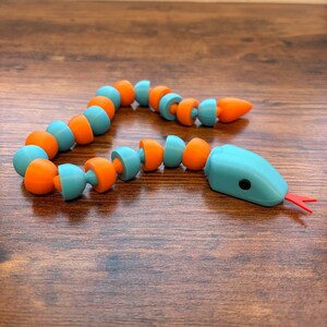 Puede incluir: Una serpiente de juguete segmentada, en colores turquesa y naranja. La cabeza es turquesa con ojos negros y una lengua bífida roja. El cuerpo está hecho de segmentos alternos turquesa y naranja, sobre una superficie de madera.