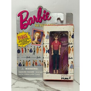 May include: A Barbie keychain doll in its original packaging. The doll wears a red and white checkered shirt and blue jeans. The packaging features the Barbie logo, illustrations of Barbie dolls, and a warning about small parts.