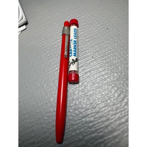 May include: A red mechanical pencil with a silver metal clip and a separate clear plastic tube containing red crayon marker leads. The tube features white labeling with blue text that reads CRAYON MARKER LEADS and the Scripto brand logo.