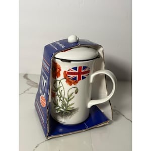 May include: White ceramic tea mug with a lid and infuser, featuring a poppy flower design. The mug is packaged in a blue box with the words "TEA MUG" and "STRAINER TEA MUG" printed on it. The mug has a handle and a "Made in England" sticker.