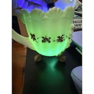 May include: A pale yellow glass pitcher with a scalloped rim and a handle, featuring a horizontal band of brown floral patterns. The pitcher is illuminated from within by a bright green light, highlighting its translucent body and three feet.