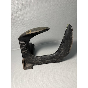 May include: A heavy, black cast iron cobbler's shoe last with a curved, upright heel section and a flat, horizontal forefoot plate. The side of the metal tool features the embossed text "NR 2" and shows signs of wear with faint yellow markings.
