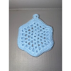 May include: A light blue hexagonal crochet coaster or ornament featuring an intricate openwork pattern with a small loop at the top for hanging. The piece is crafted from yarn with a consistent stitch structure throughout the entire design.