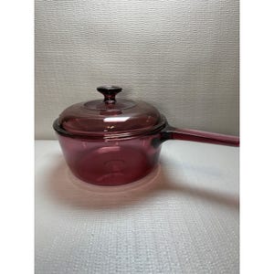 May include: A vintage, ruby-colored glass saucepan with a matching lid. The saucepan has a long handle and is designed for stovetop cooking. The glass is transparent, allowing visibility of the contents. The lid has a knob for easy handling.