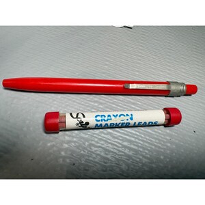 May include: A red Scripto mechanical pencil and a tube of crayon marker leads. The pencil is a solid red color with a silver metal clip and the word "Scripto" printed on the side. The tube of leads is white with blue text that reads "Crayon Marker Leads".