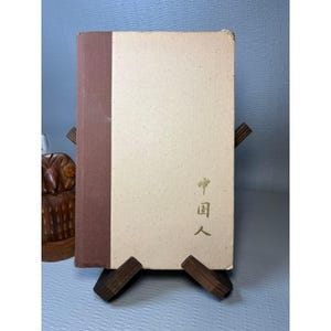 May include: A hardcover book with a brown spine and a cream-colored cover. The front cover features gold-colored Japanese characters vertically aligned on the right side. The book is displayed on a dark wooden stand, showing a clean, rectangular design.