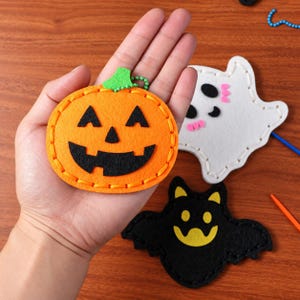 Op de afbeelding: Een oranje vilt pompoenornament met een zwart vilt jack-o'-lantern gezicht en groene steel, naast een wit vilt spookornament met zwarte ogen en roze wangen, en een zwart vilt vleermuisornament met gele ogen en een gele mond.