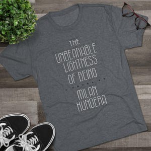 May include: A grey t-shirt featuring the text "The Unbearable Lightness of Being" by Milan Kundera in white. The shirt is displayed with black and white trainers and spectacles on a wooden surface.