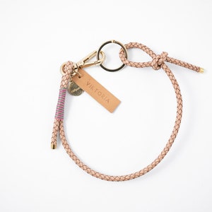 Op de afbeelding: Een lichtbruine leren sleutelhanger met een gouden metalen ring en sluiting. De sleutelhanger heeft een gevlochten leren band met een roze en witte gestreepte accent. De sleutelhanger heeft ook een klein leren label met het woord "Viktoria" erop gedrukt.