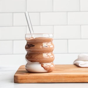 May include: A clear, rippled glass tumbler filled with a thick, brown chocolate smoothie or mousse. A straight, transparent glass straw is inserted into the center of the drink. The glass features three distinct horizontal ridges along its body.