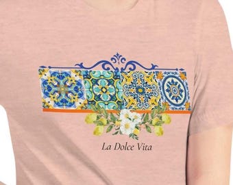 May include: A light pink t-shirt featuring a decorative design with four square panels of blue and yellow tile patterns. Below the tiles are white flowers and the text "La Dolce Vita".