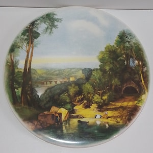 May include: A circular decorative plate with a landscape painting of a bridge, trees, and water. The plate has a white edge and a painted image in green, blue, and brown tones. The scene shows a tranquil outdoor environment.