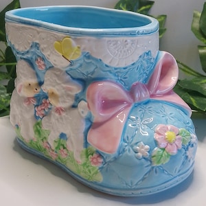 May include: A ceramic boot-shaped planter in shades of blue, white, and pink. The planter features a bow, lamb figures, and floral accents. The interior is light blue, and the exterior has a quilted pattern.